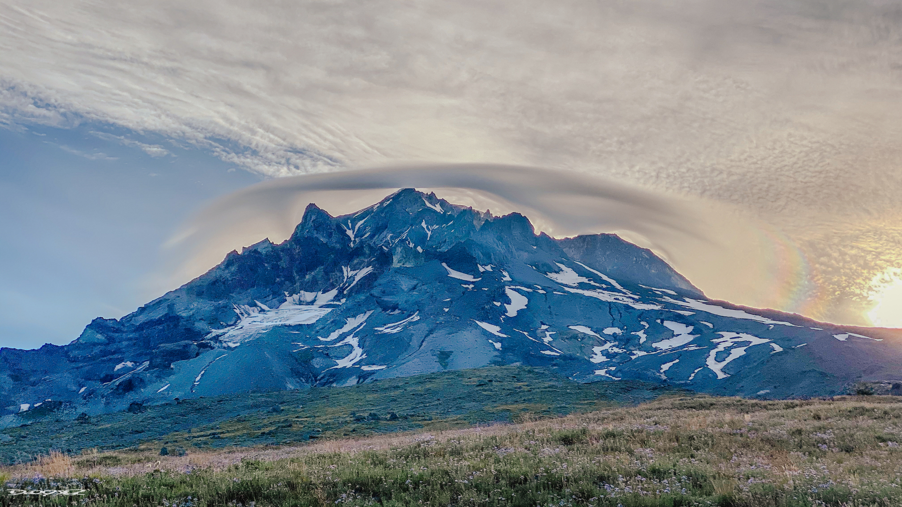 A dramatic landscape features a rugged Wy'East with swirling clouds above and patches of snow.