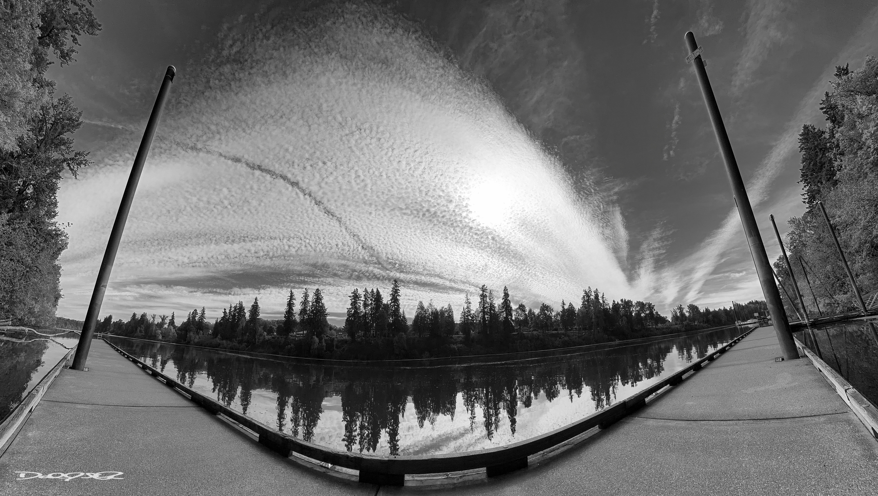 A black and white photograph of a serene Willamette River is framed by two dock piers, with a dramatic sky featuring streaky clouds overhead.