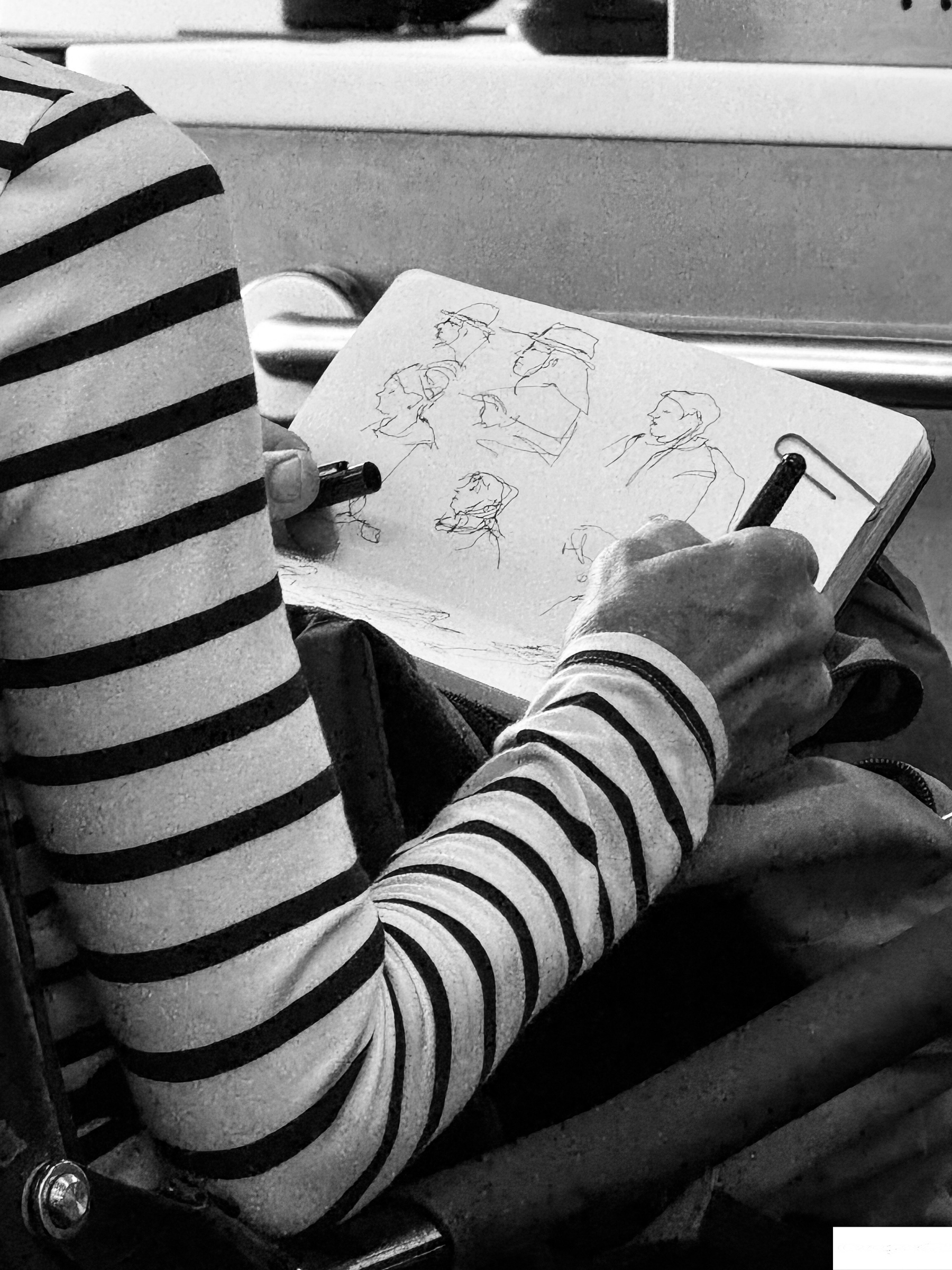 A person wearing a striped shirt is sketching various face profiles in a notebook.