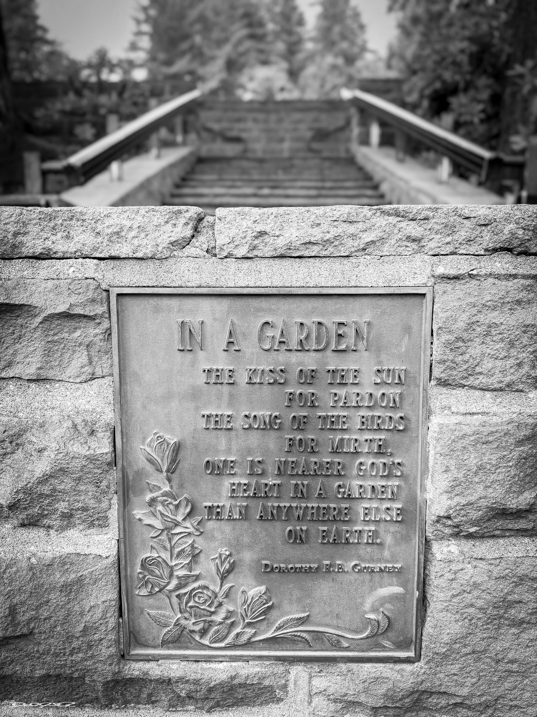 A plaque mounted on a stone wall, engraved with a nature-themed poem by Dorothy Frances Gurney, is seen in focus with a blurred garden pathway in the background.