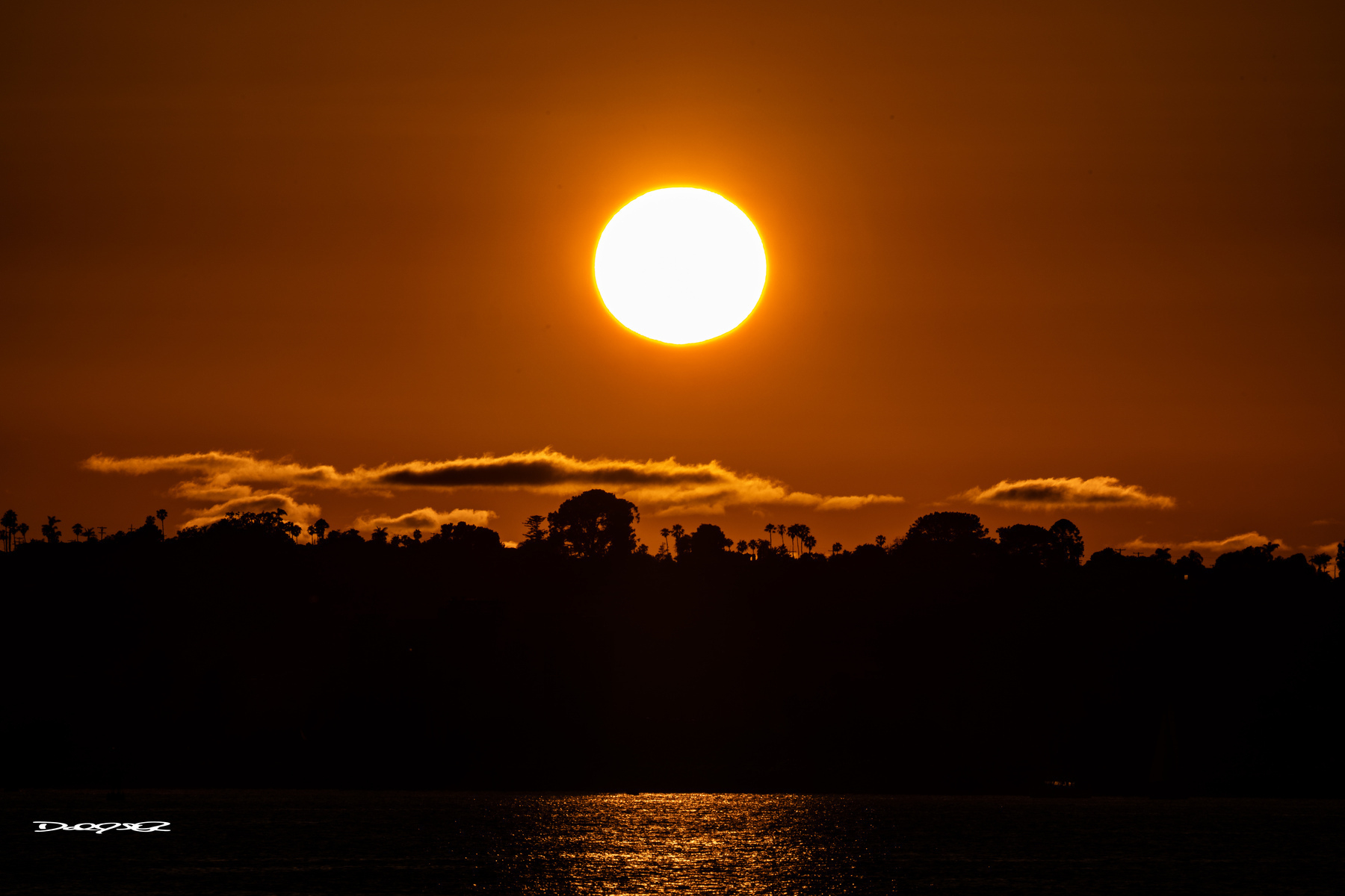 A bright sun sets over a silhouetted landscape with a reflective body of water in the foreground.
