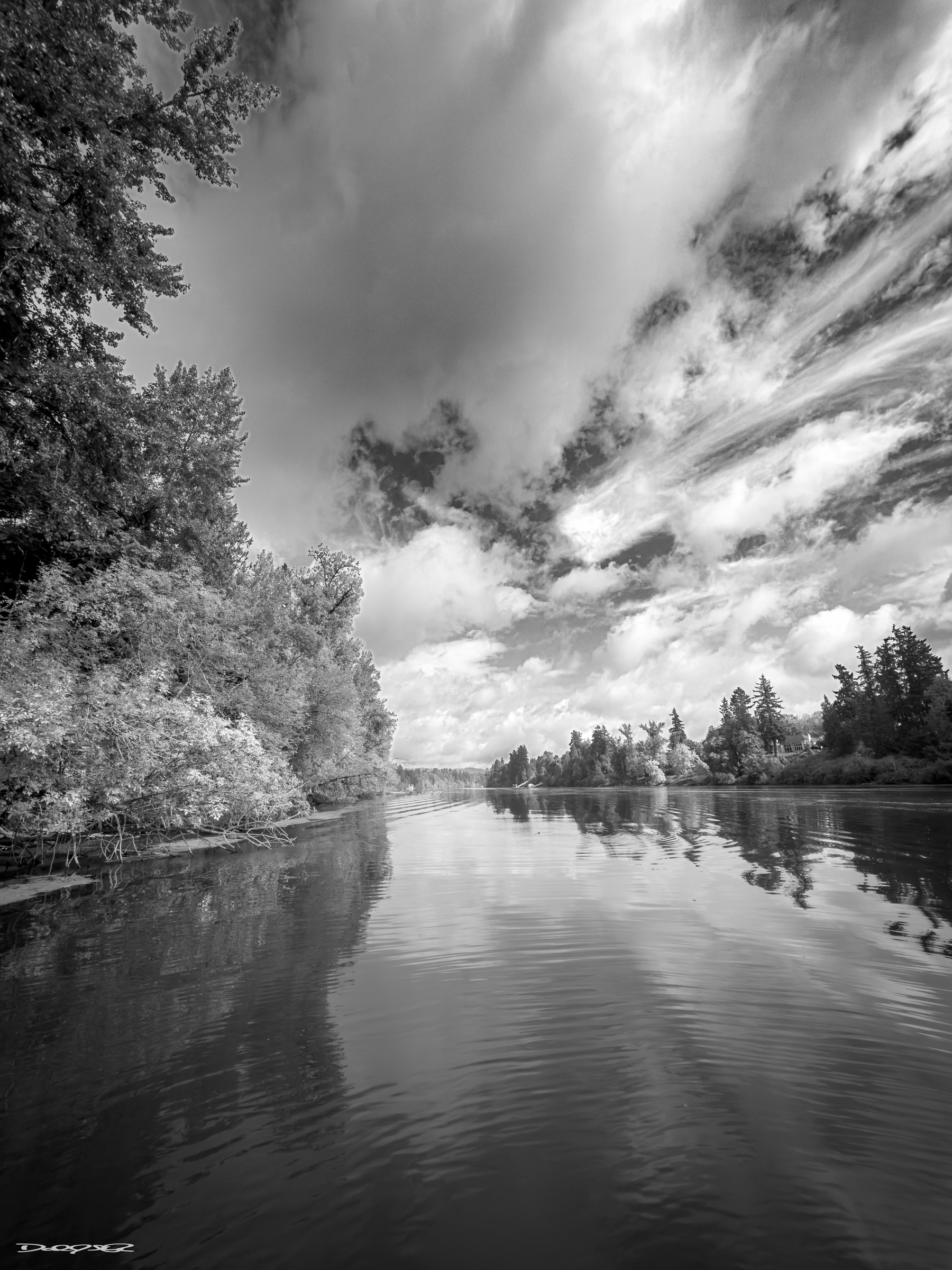 A tranquil river scene is captured in black and white, with trees lining the banks under a dramatic, cloud-filled sky.