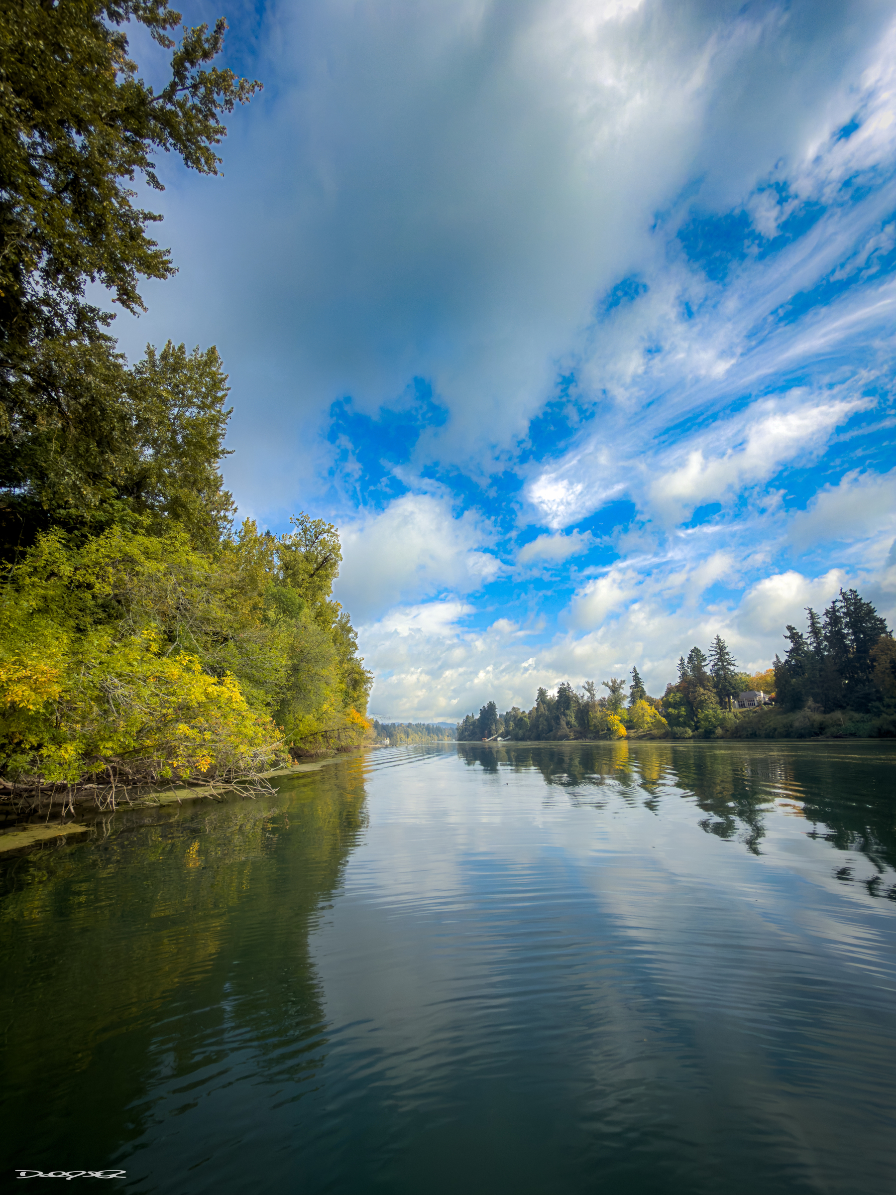 A serene river scene features lush trees on both sides and a partly cloudy blue sky reflecting on the water's surface.